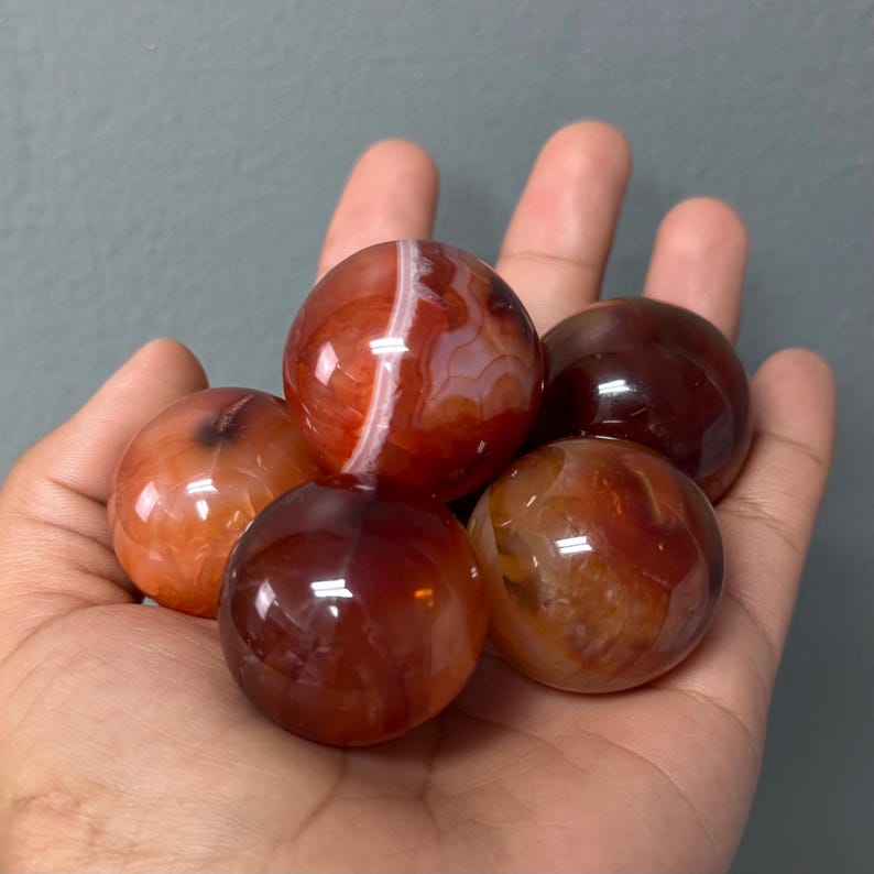 May include: Five polished carnelian spheres in shades of red, orange, and brown are held in a hand. The spheres have a smooth, reflective surface with natural banding patterns. The gemstones vary in colour and pattern.