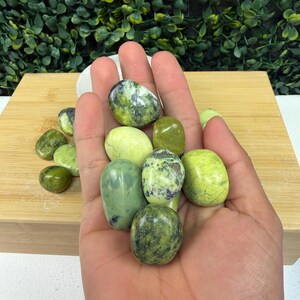 May include: A hand holding a collection of polished green stones. The stones vary in shades of green, from light to dark, with some displaying speckled patterns. The stones are smooth and oval-shaped, suggesting they are tumbled or polished.