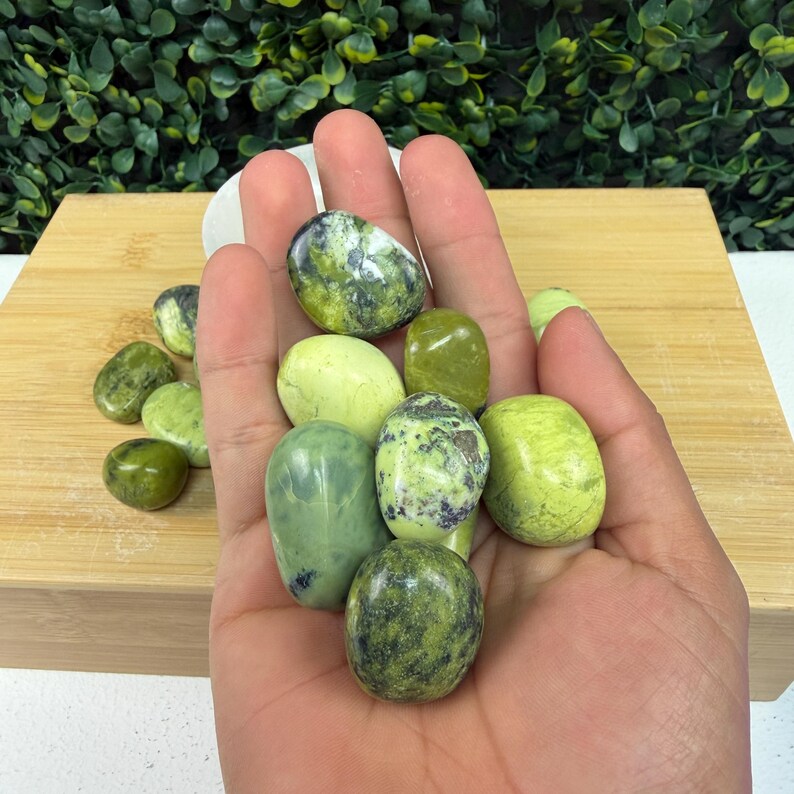 May include: A hand holding a collection of polished green stones. The stones vary in shades of green, from light to dark, with some displaying speckled patterns. The stones are smooth and oval-shaped, suggesting they are tumbled or polished.