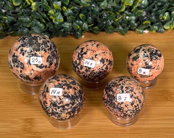 May include: Five polished spherical stones with a speckled pink, orange, and black granite-like pattern. Each sphere rests on a clear acrylic stand and features a small white label with text reading 25G, 14H, 14I, 8J, and 8K respectively.