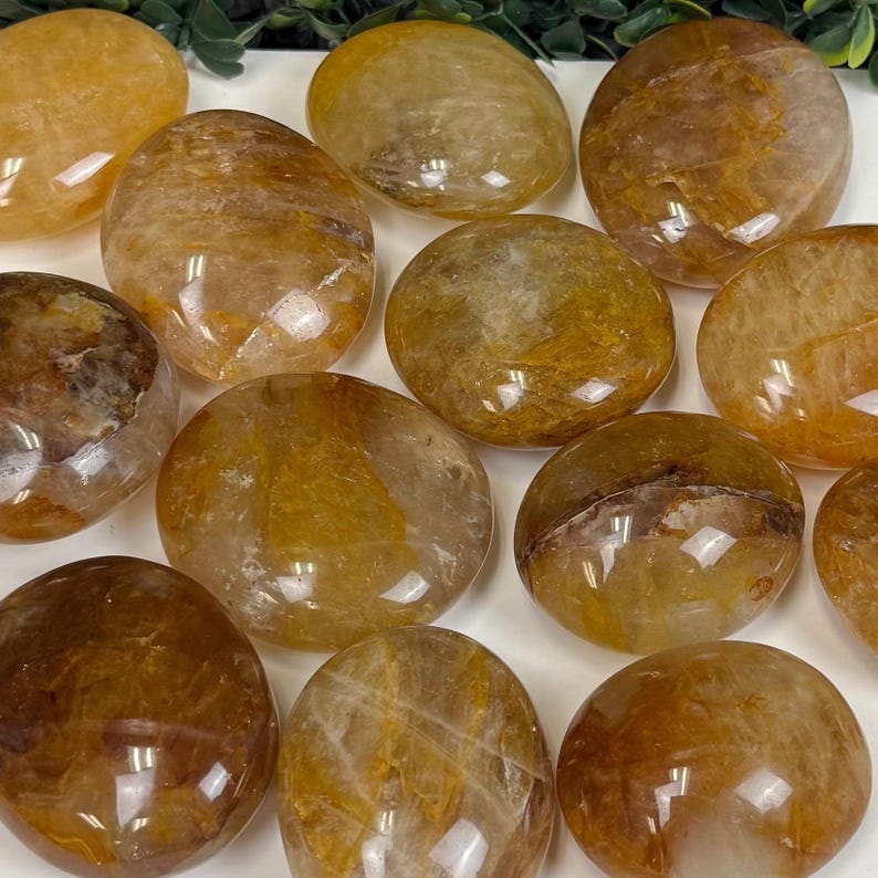 May include: A collection of polished, oval-shaped golden rutile quartz stones. The stones vary in shades of yellow and brown, with internal veining and a glossy finish. These natural gemstones are ideal for crystal healing and meditation.