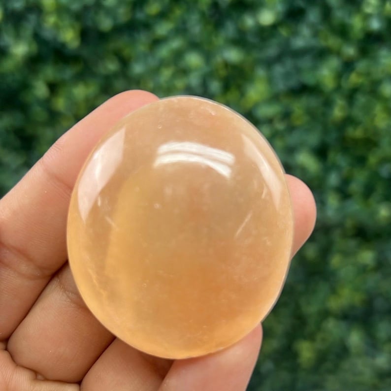Pode incluir: Uma pedra preciosa lisa, de forma oval, amarelo-alaranjada, &eacute; segurada em uma m&atilde;o. A pedra polida tem uma superf&iacute;cie brilhante, refletindo a luz. O fundo &eacute; verde desfocado, sugerindo folhagem. Esta pedra preciosa &eacute; provavelmente usada para fins de cura ou decora&ccedil;&atilde;o.