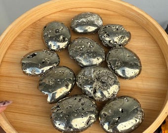 May include: A collection of nine polished pyrite stones with a metallic gold luster and irregular, textured surfaces. Each stone features a unique, rounded shape with natural indentations and crystalline facets, displayed together on a round wooden tray.