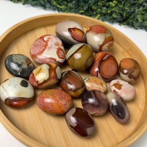 Puede incluir: Una colección de piedras pulidas en varios colores, incluyendo marrón, rojo, crema y negro, dispuestas en una bandeja redonda de madera. Las piedras tienen patrones y texturas únicos, algunas lisas y otras más rugosas.