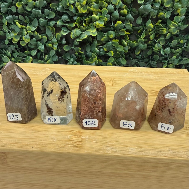 May include: Five quartz crystal points in shades of brown and clear are displayed on a wooden surface. Each crystal has a white label with a number and letter. The crystals vary in size and have inclusions.