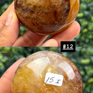 May include: Two polished, spherical gemstones with a mix of golden, brown, and translucent hues. The top stone has the text "#12" in a black rectangle. The bottom stone has the text "15 I" on a white rectangle. The stones are held in a hand.