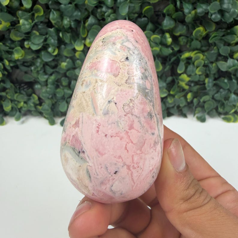 May include: A pink and white gemstone egg with a marbled appearance. The stone has a smooth, polished surface and is held in a hand. The background is a green hedge.