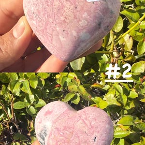 May include: Two heart-shaped stones with pink and gray marbling are held up against a backdrop of green foliage. One stone has the number "34A" on a white sticker. The other stone has the number "#2" on a white sticker.