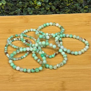 May include: Several bracelets made of small, round, green and brown beads. The bracelets are arranged in a circular shape, with varying sizes and bead colors. The beads have a natural, earthy appearance, and the bracelets are displayed on a wooden surface.