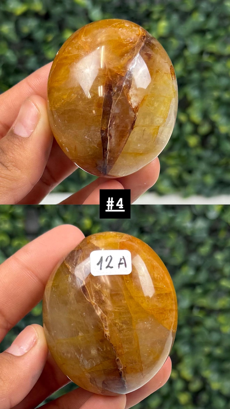 May include: Two polished, golden-brown quartz spheres. The spheres have internal veining and inclusions, with the text "#4" and "12 A" visible. The spheres are held in a hand against a blurred green background.
