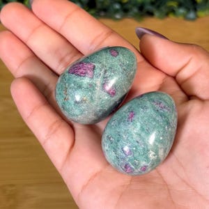 May include: Two polished, egg-shaped stones featuring a mottled green base color with embedded deep magenta and dark red crystalline inclusions. The surfaces are smooth and reflective, showcasing a natural mineral texture throughout.