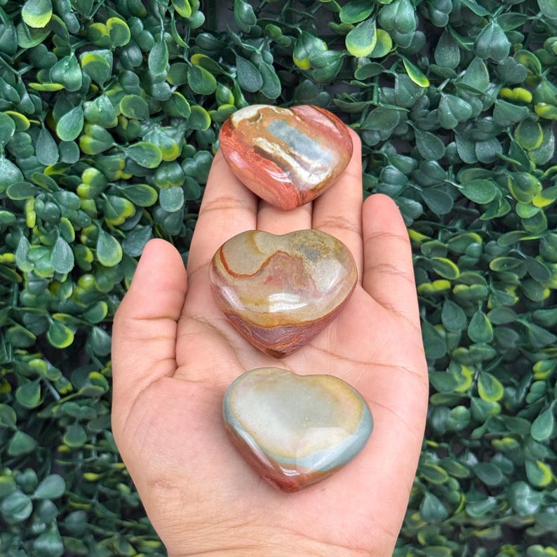 Puede incluir: Se muestran tres piedras en forma de coraz&oacute;n en tonos rojos, marrones y grises. Las piedras tienen una superficie pulida y lisa y se sostienen en una mano sobre un fondo de follaje verde. Las piedras miden aproximadamente 5 cm de largo.