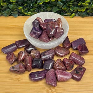 May include: A collection of polished, tumbled gemstones in shades of deep purple and burgundy. The stones are displayed in a white, shallow bowl and scattered on a wooden surface. The gemstones vary in size and shape, with a glossy finish.