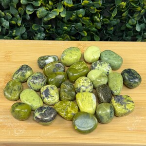 May include: A collection of tumbled green stones with varying shades and patterns. The stones range from light to dark green, some with black or gray inclusions. They are arranged on a light brown wooden surface.
