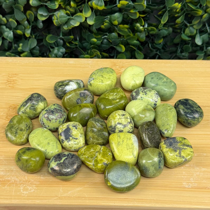 May include: A collection of tumbled green stones with varying shades and patterns. The stones range from light to dark green, some with black or gray inclusions. They are arranged on a light brown wooden surface.