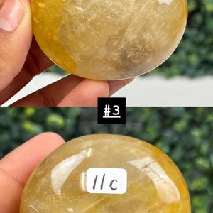 May include: Two golden-yellow, polished crystal spheres. The top sphere has a smooth, rounded shape. The bottom sphere has a white label with the text "11c" and the number "3" in a black box. The crystals are held in a hand.