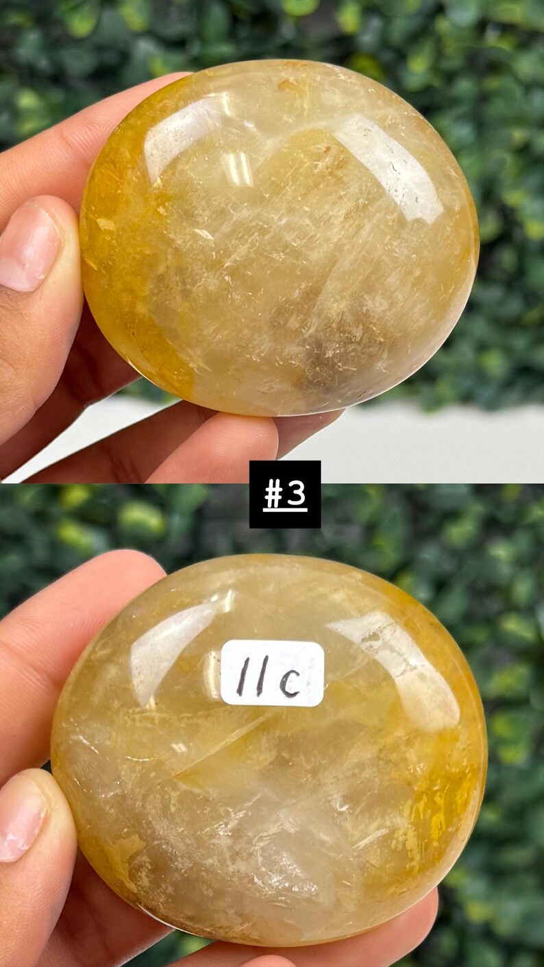 May include: Two golden-yellow, polished crystal spheres. The top sphere has a smooth, rounded shape. The bottom sphere has a white label with the text "11c" and the number "3" in a black box. The crystals are held in a hand.