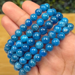 May include: A collection of vibrant blue apatite gemstone bead bracelets. The beads are round and have a polished, glossy finish. The bracelets are held in a hand, showcasing the color and texture of the stones. The bracelets are a beautiful accessory.