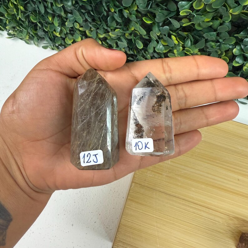 May include: Two clear quartz crystal towers with brown inclusions. The taller tower has the number "12J" on a white sticker, and the shorter tower has "10K" on a white sticker. The crystals are held in a hand.
