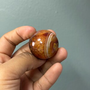 May include: A polished, spherical gemstone with a warm colour palette. The orb features bands of orange, brown, and white, creating a marbled effect. The stone is held in a hand, showcasing its smooth, reflective surface. The gemstone is approximately 5 cm in diameter.