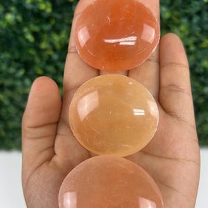 Pode incluir: Tr&ecirc;s pedras polidas de cor laranja s&atilde;o seguradas em uma m&atilde;o. As pedras, em forma oval, t&ecirc;m uma superf&iacute;cie lisa e brilhante. O fundo &eacute; um verde desfocado.