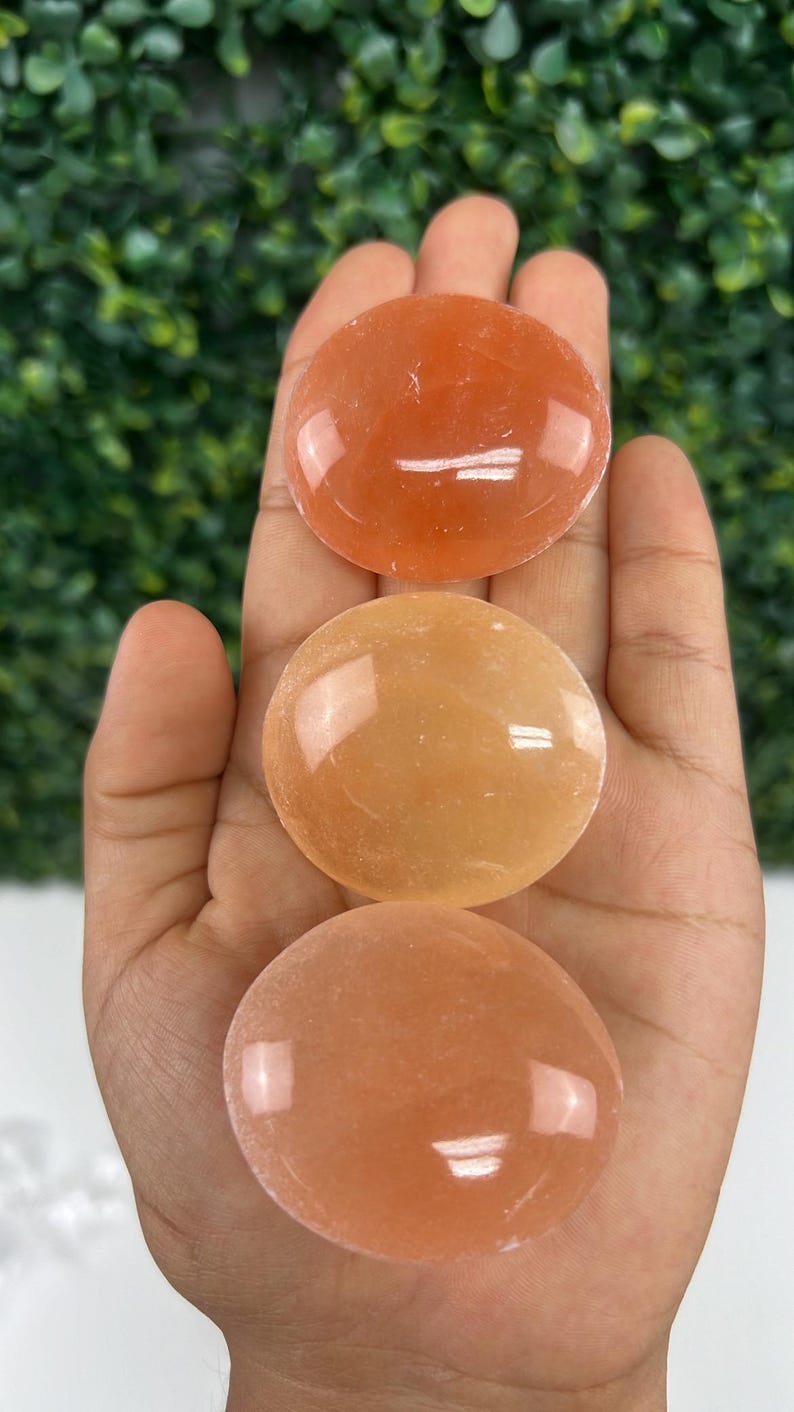Pode incluir: Tr&ecirc;s pedras polidas de cor laranja s&atilde;o seguradas em uma m&atilde;o. As pedras, em forma oval, t&ecirc;m uma superf&iacute;cie lisa e brilhante. O fundo &eacute; um verde desfocado.