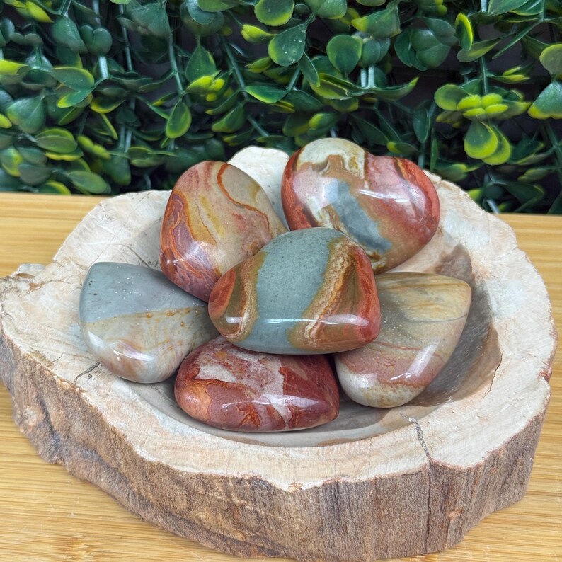 Puede incluir: Una colecci&oacute;n de piedras pulidas en tonos rojos, marrones, beiges y grises, expuestas en un cuenco de madera r&uacute;stica. Las piedras tienen una forma lisa y redondeada y un aspecto veteado. El cuenco es de madera clara.