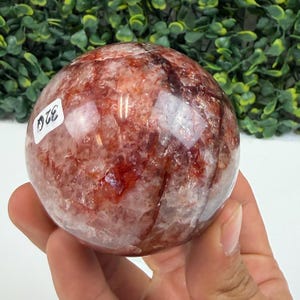 May include: A polished, spherical gemstone with a mix of red and clear quartz. The stone has a marbled appearance with visible inclusions and a label with the number "320". The sphere is held in a hand.