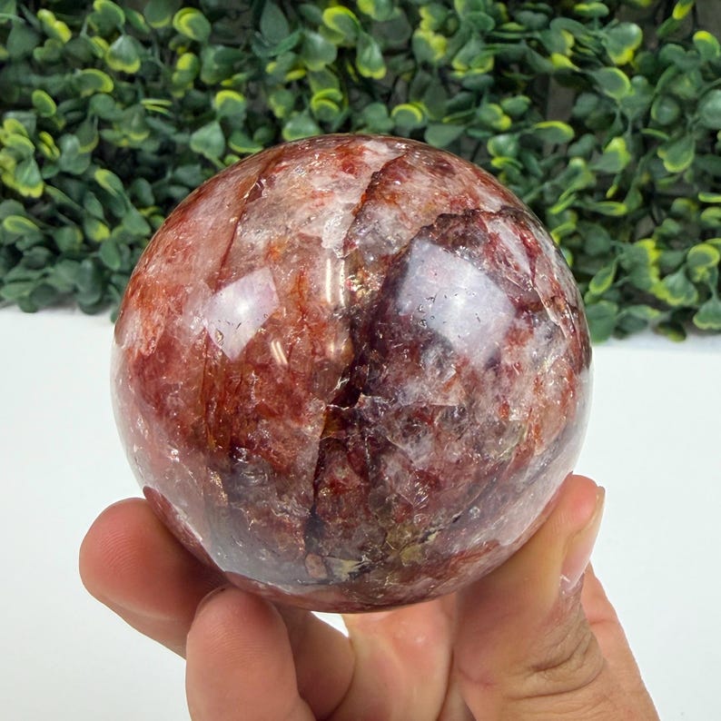May include: A polished, spherical gemstone with a mix of red, white, and clear inclusions. The orb is held in a hand, showcasing its smooth, reflective surface. The background features green foliage.