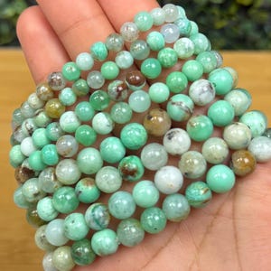 May include: A collection of green and white gemstone bead bracelets. The beads vary in shades of green, with some displaying brown and white patterns. The bracelets are displayed on a hand, showcasing the natural variations in the stones.