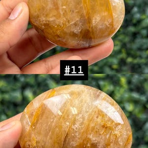 May include: Two golden-brown, polished gemstones are held in a person's hand. The stones have a smooth, rounded shape with internal striations. One stone has a sticker that says "146" and the other has a sticker that says "#11".