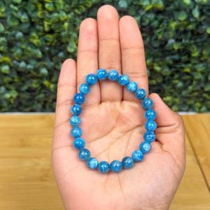 May include: A blue beaded bracelet with a stretch band. The bracelet is made of round, light blue beads. The bracelet is shown on a person's hand.