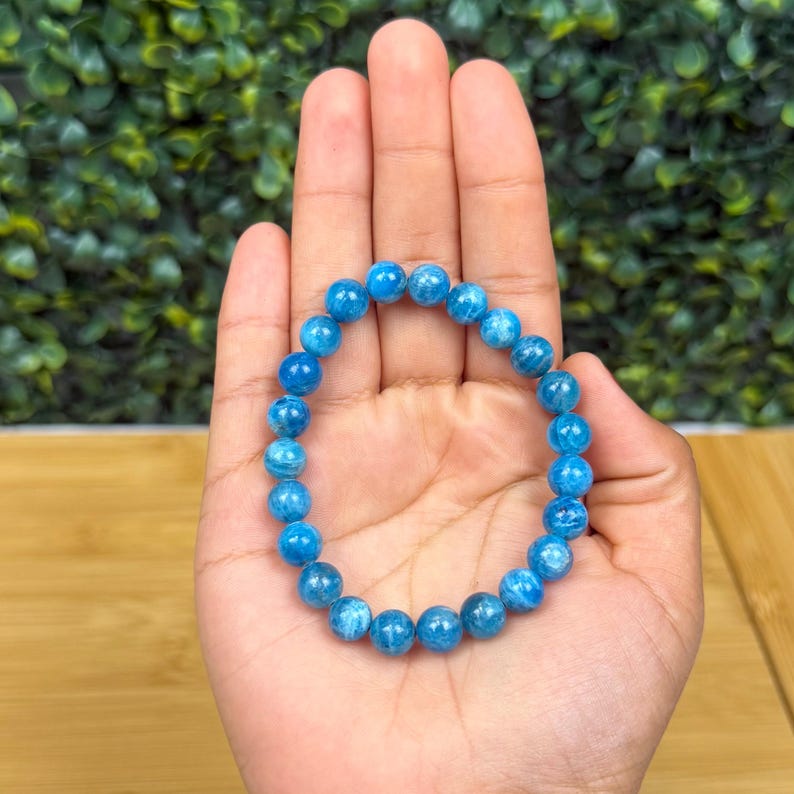 May include: A blue beaded bracelet with a stretch band. The bracelet is made of round, light blue beads. The bracelet is shown on a person's hand.