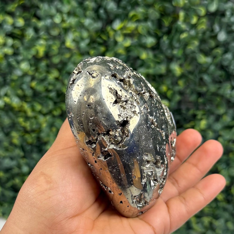 May include: A polished, metallic pyrite stone, also known as fool's gold, with a rough, textured surface. The stone is held in a hand, showcasing its reflective, silver-like appearance. The background is a blurred green.