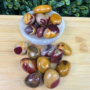 May include: A collection of polished stones in shades of red, yellow, and brown, some with a marbled appearance. The stones are displayed in a white bowl and scattered on a wooden surface. The stones vary in size and shape.