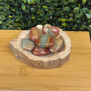 Puede incluir: Una colecci&oacute;n de piedras pulidas en tonos rojos, marrones y verdes, colocadas en un cuenco de madera r&uacute;stico. El cuenco est&aacute; hecho de una secci&oacute;n de tronco de &aacute;rbol, agregando un elemento natural. Las piedras tienen un acabado liso y brillante.