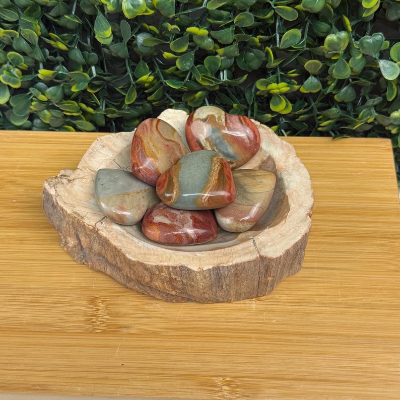 Puede incluir: Una colecci&oacute;n de piedras pulidas en tonos rojos, marrones y verdes, colocadas en un cuenco de madera r&uacute;stico. El cuenco est&aacute; hecho de una secci&oacute;n de tronco de &aacute;rbol, agregando un elemento natural. Las piedras tienen un acabado liso y brillante.