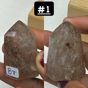 May include: Two clear quartz crystal points with inclusions, one labeled "#1" and the other with "8T". The crystals are held in a person's hands. The crystals have a pointed top and a faceted body.