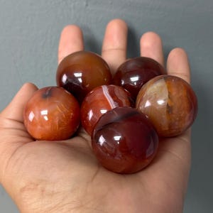 May include: Six polished carnelian spheres in shades of red, orange, and brown are displayed. The spheres have a smooth, glossy surface and vary in colour and pattern. The stones are held in a hand against a grey background.