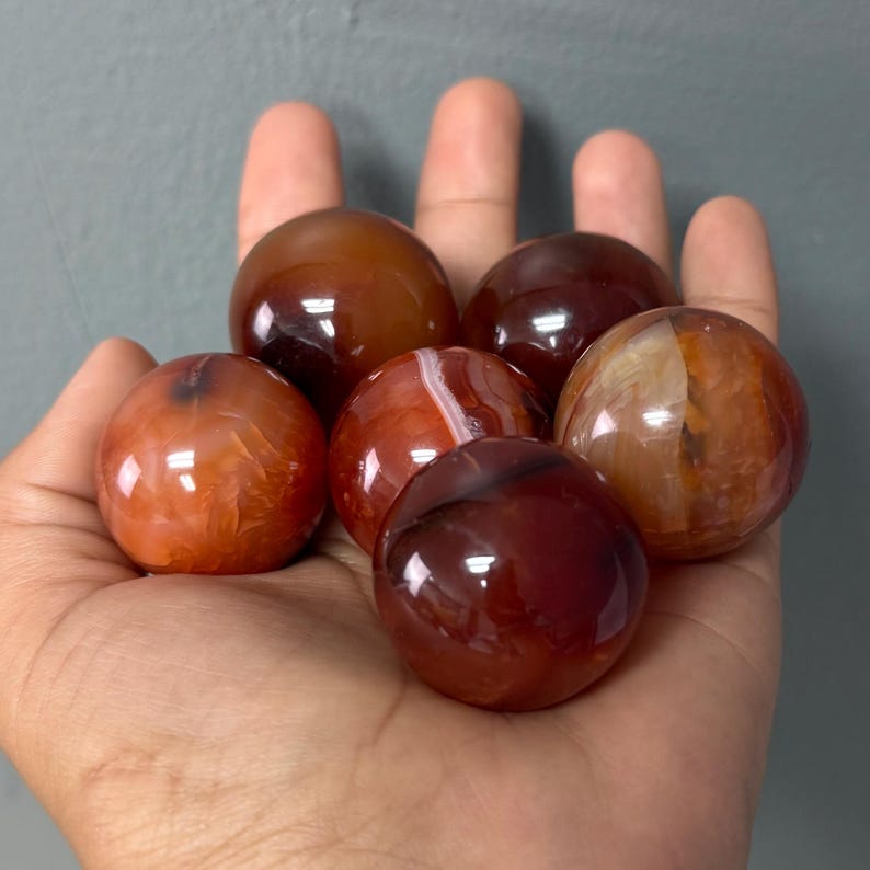 May include: Six polished carnelian spheres in shades of red, orange, and brown are displayed. The spheres have a smooth, glossy surface and vary in colour and pattern. The stones are held in a hand against a grey background.