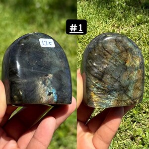 May include: A polished labradorite stone with a domed shape, displaying iridescent flashes of blue, gold, and green against a dark gray base. The stone features a white sticker labeled 13c and is marked with the text #1 in a black box.