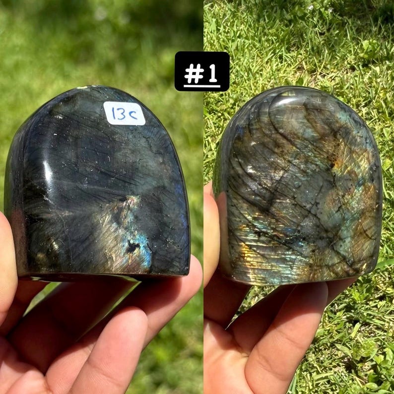 May include: A polished labradorite stone with a domed shape, displaying iridescent flashes of blue, gold, and green against a dark gray base. The stone features a white sticker labeled 13c and is marked with the text #1 in a black box.