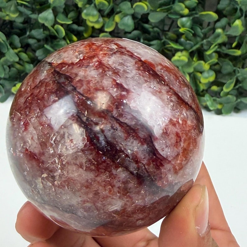 May include: A clear, spherical crystal ball with red and white veining. The polished gemstone is held in a hand, showcasing its natural patterns and textures. The crystal ball is a decorative item.