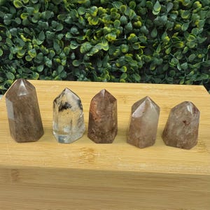 May include: Five polished quartz crystal points in shades of brown and clear. Each crystal has a flat base and a pointed top. The crystals are arranged on a light brown wooden surface, with a backdrop of green foliage.