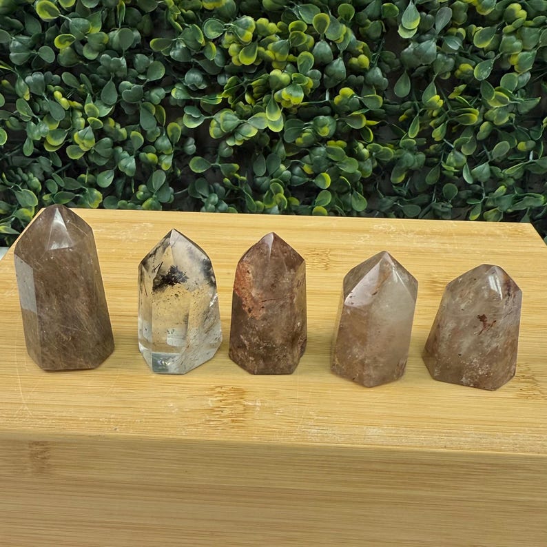 May include: Five polished quartz crystal points in shades of brown and clear. Each crystal has a flat base and a pointed top. The crystals are arranged on a light brown wooden surface, with a backdrop of green foliage.