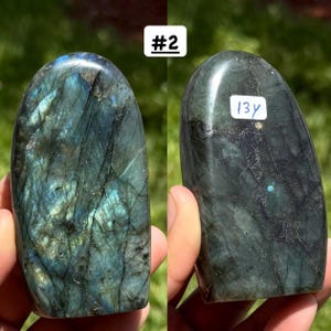 May include: A polished labradorite stone with a rounded top, featuring dark grey and black tones with iridescent blue and green flashes. The stone displays natural veining and fractures. A white sticker with the handwritten text 13y is attached.