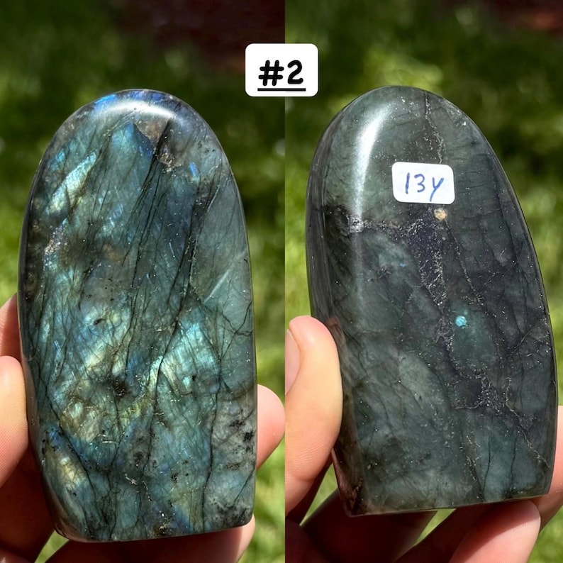 May include: A polished labradorite stone with a rounded top, featuring dark grey and black tones with iridescent blue and green flashes. The stone displays natural veining and fractures. A white sticker with the handwritten text 13y is attached.