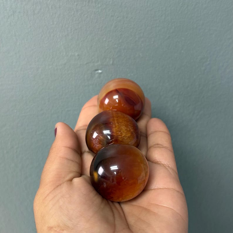 May include: Three polished gemstone spheres in shades of brown and orange. The spheres are smooth and reflective, with variations in colour and pattern. The gemstones are held in a hand against a grey background.