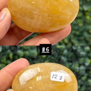 May include: A smooth, oval-shaped, golden-yellow gemstone. The stone is held in a hand, with the text "#6" and "12 z" visible on the stone. The background is a blurred green.
