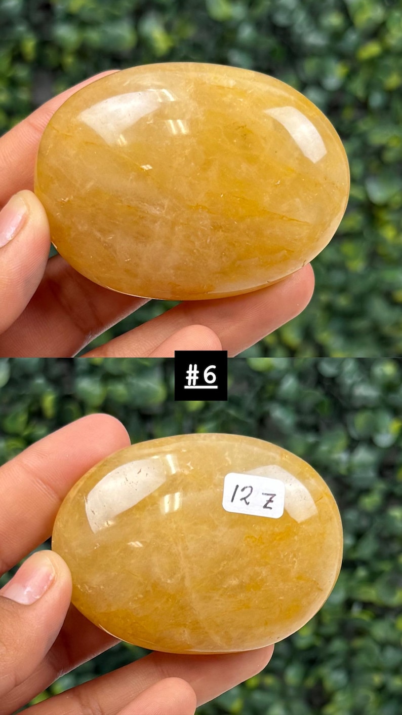 May include: A smooth, oval-shaped, golden-yellow gemstone. The stone is held in a hand, with the text "#6" and "12 z" visible on the stone. The background is a blurred green.